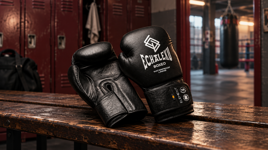 Guantes ELEMENT negros y plata de ECHALEKU BOXEO sobre un banco de vestuario recreado, con taquillas y un saco al fondo.